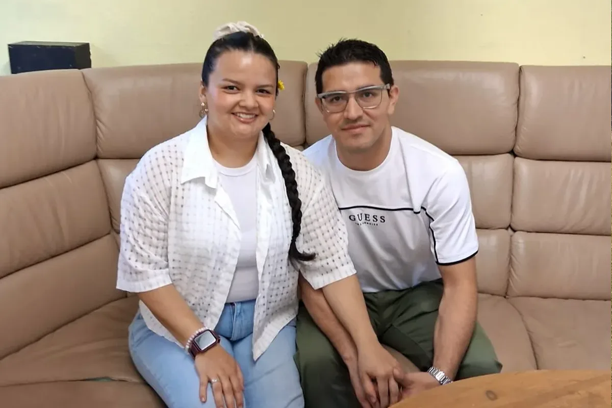 Two adults sit close together on a tan sectional couch, smiling at the camera; woman with long braid wearing white shirt and jeans, man with glasses in a white GUESS shirt and green pants.