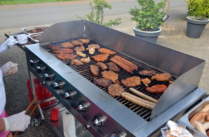 Outdoor gas grill filled with burgers, sausages, and meat grilling; cook wearing gloves stands nearby to tend the grill.