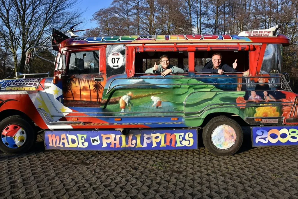 Bright, mural-painted jeepney with two men leaning from windows and giving thumbs up; 'Made in Philippines' sign visible along the side