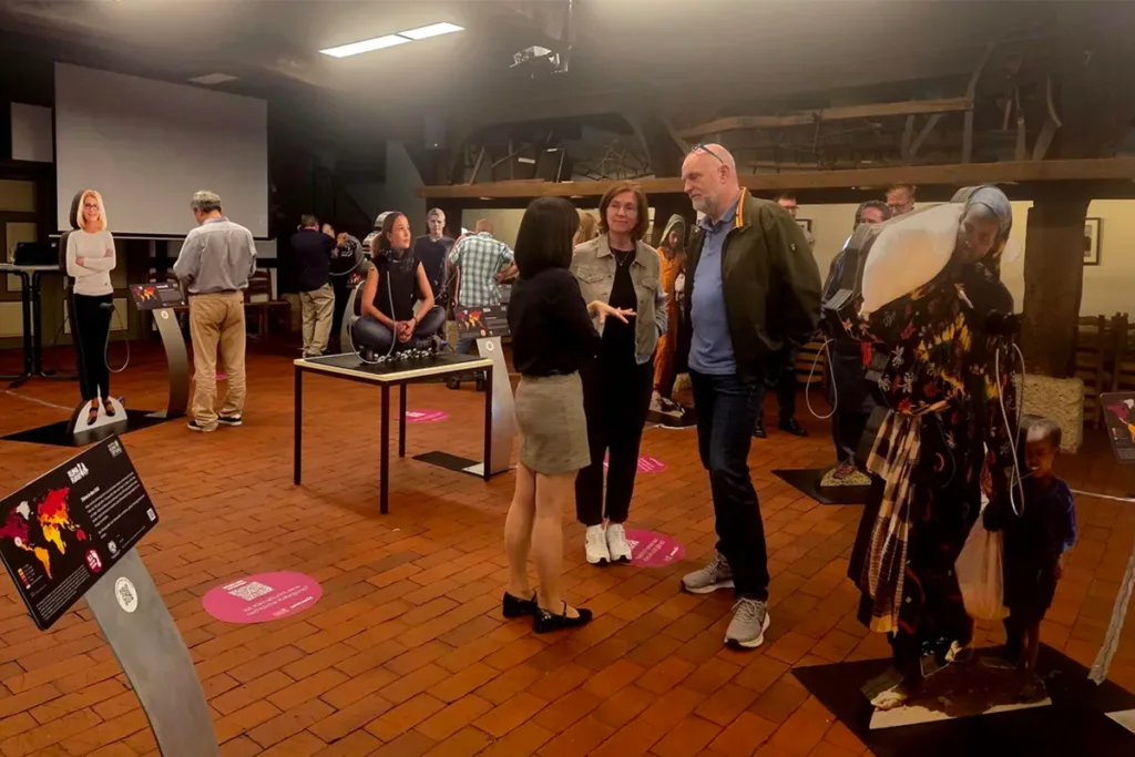Group of adults conversing and exploring an indoor exhibit with display stands and informational plaques on a brick floor.