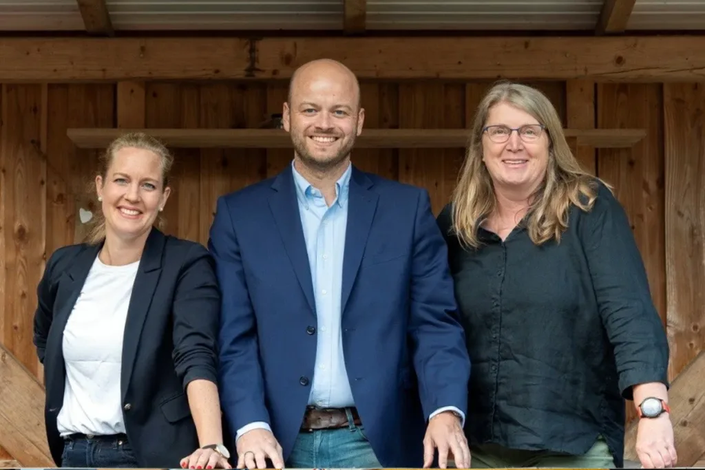 Three professionals pose together in front of a wooden backdrop, smiling at the camera.