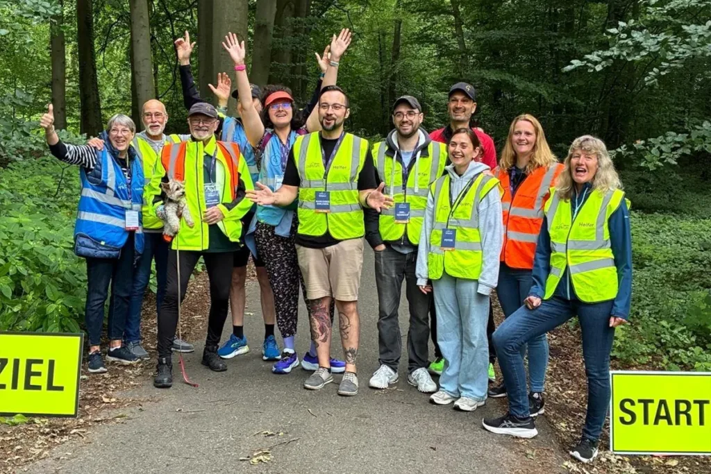 Group of volunteers in high-visibility vests posing together on a forest trail at a start line, smiling and raising their hands to celebrate.