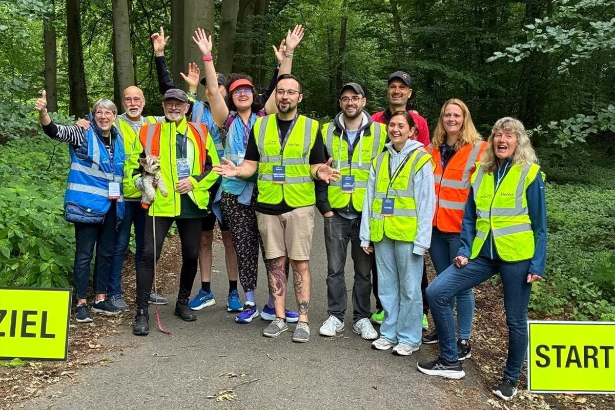 Group of volunteers in high-visibility vests posing together on a forest trail at a start line, smiling and raising their hands to celebrate.