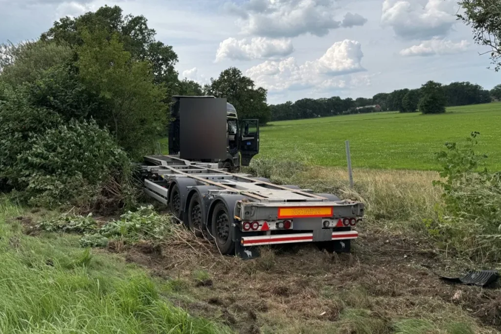 Truck trailer stuck in a muddy ditch by the roadside, with the cab visible in the background on a rural field.