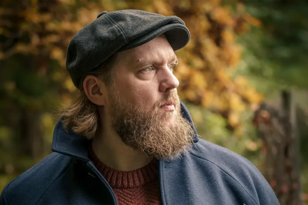 Bearded man wearing a dark cap and navy jacket outdoors in autumn foliage, looking to the right.
