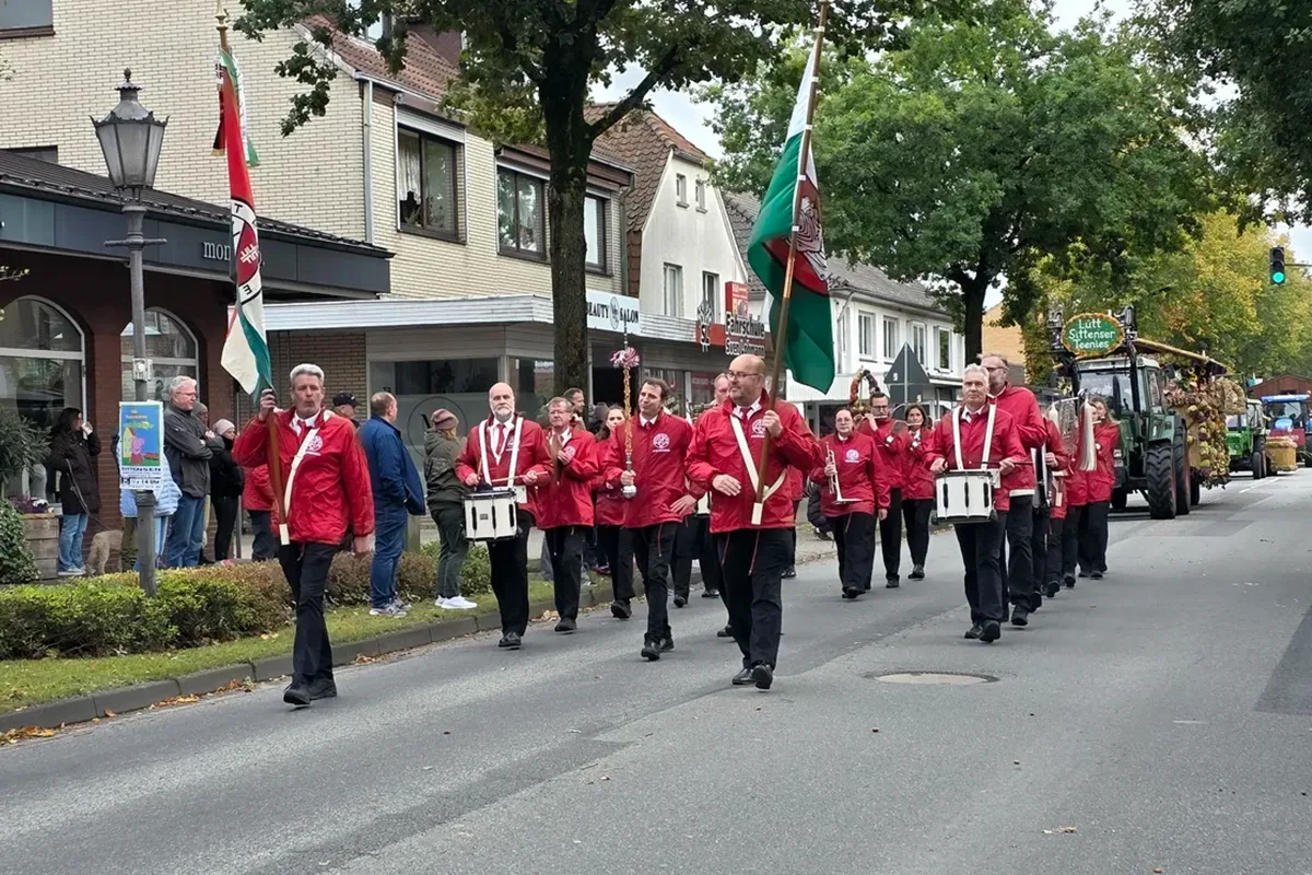 Parade marching band in red jackets marching down a street with flags and drummers, onlookers on the sidewalk.