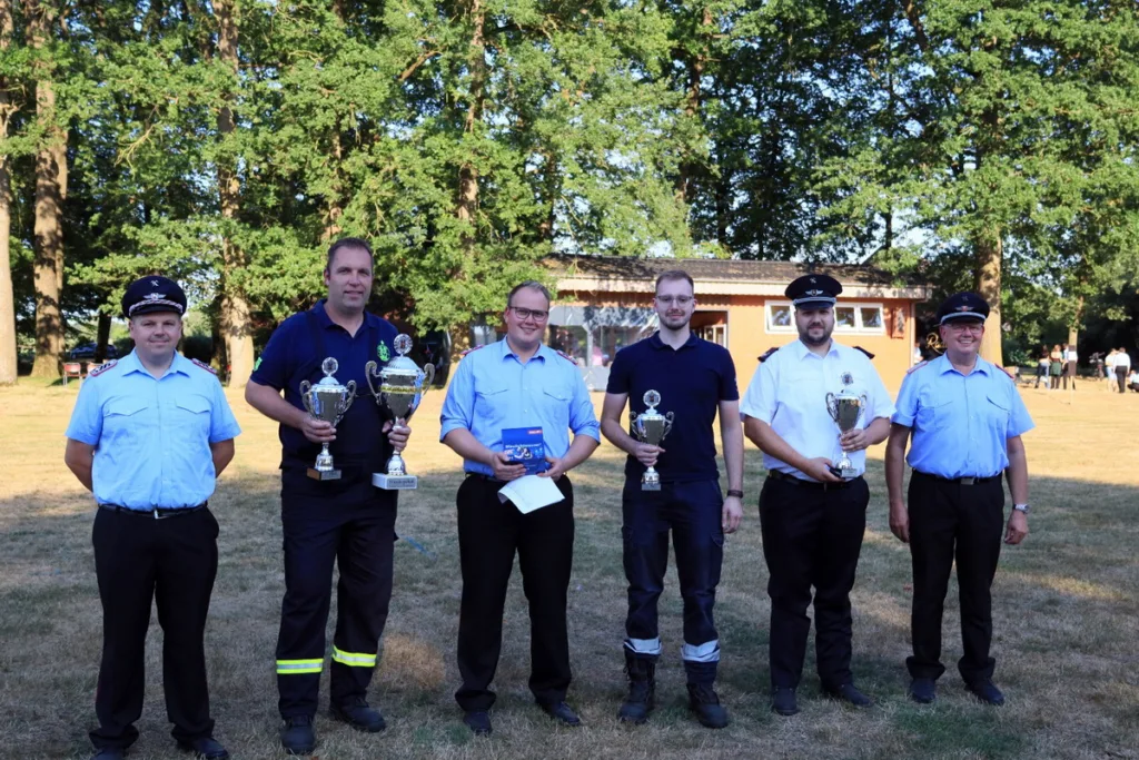 Six uniformed men standing on a grassy field; several hold trophies with a building and trees behind them in sunlight on a clear day.