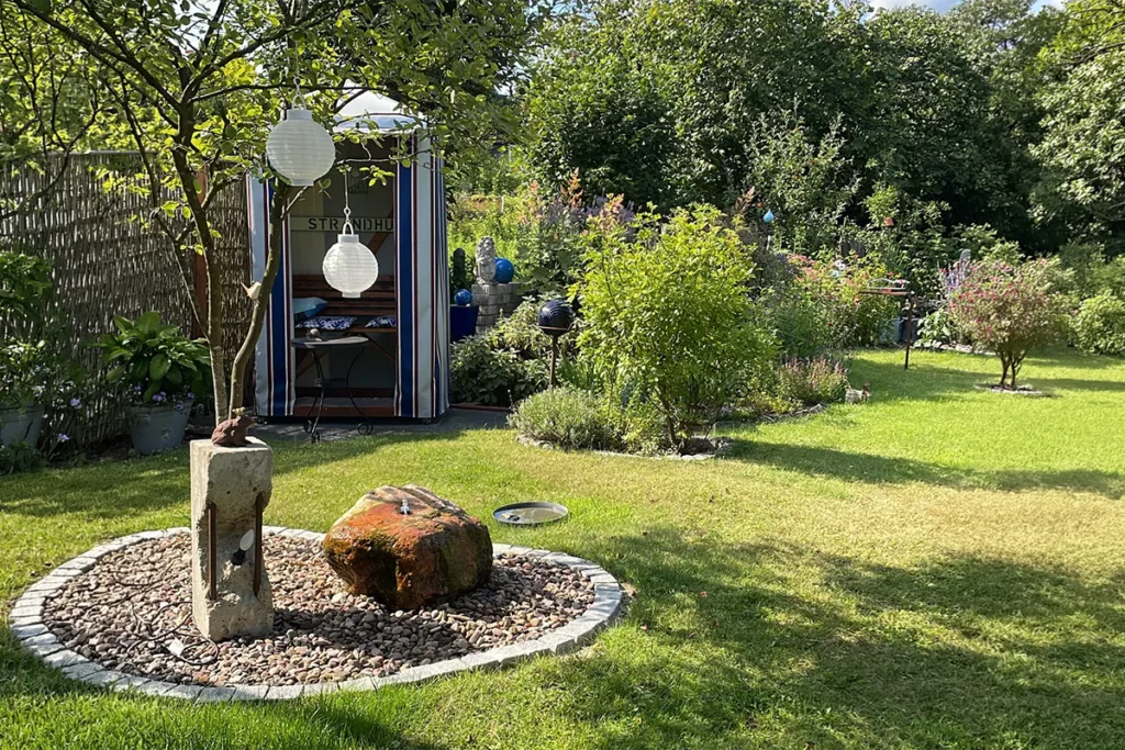 Sunlit backyard garden with a striped blue-and-white shed, paper lanterns hanging from a tree, and a circular pebble bed with a stone sculpture in the foreground