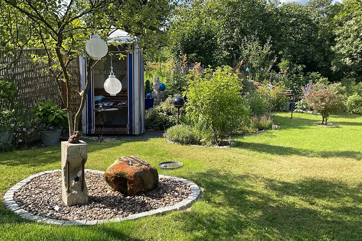 Sunlit backyard garden with a striped blue-and-white shed, paper lanterns hanging from a tree, and a circular pebble bed with a stone sculpture in the foreground