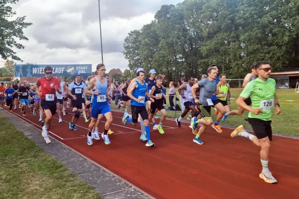 Group of runners starting a race on a red track, wearing numbered bibs and colorful athletic gear, with a blue banner in the background saying 'START/ZIEL'.