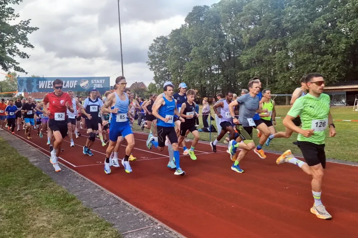Group of runners starting a race on a red track, wearing numbered bibs and colorful athletic gear, with a blue banner in the background saying 'START/ZIEL'.