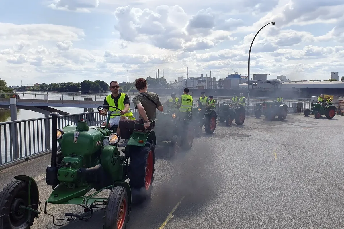 Line of vintage green tractors with drivers in high-visibility vests on a bridge, smoke billowing from the engines as they move along.