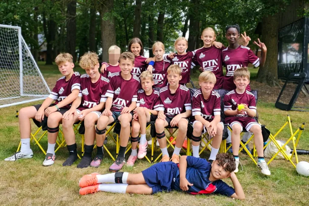 Group of young soccer players in maroon uniforms pose for a team photo outdoors, with a goal and trees in the background and one boy lying on the grass in front.