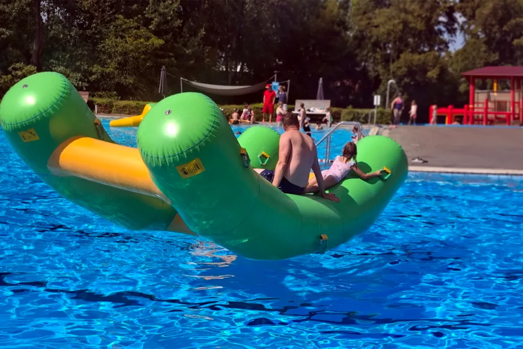 Two people sit on a large green inflatable banana boat floating in a bright blue pool with a pool deck in the background.