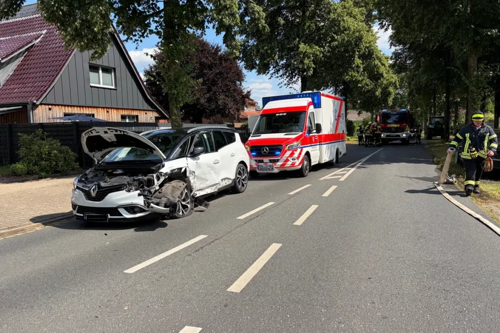 Two damaged cars after a collision on a residential street, with an ambulance and firefighters in the background.