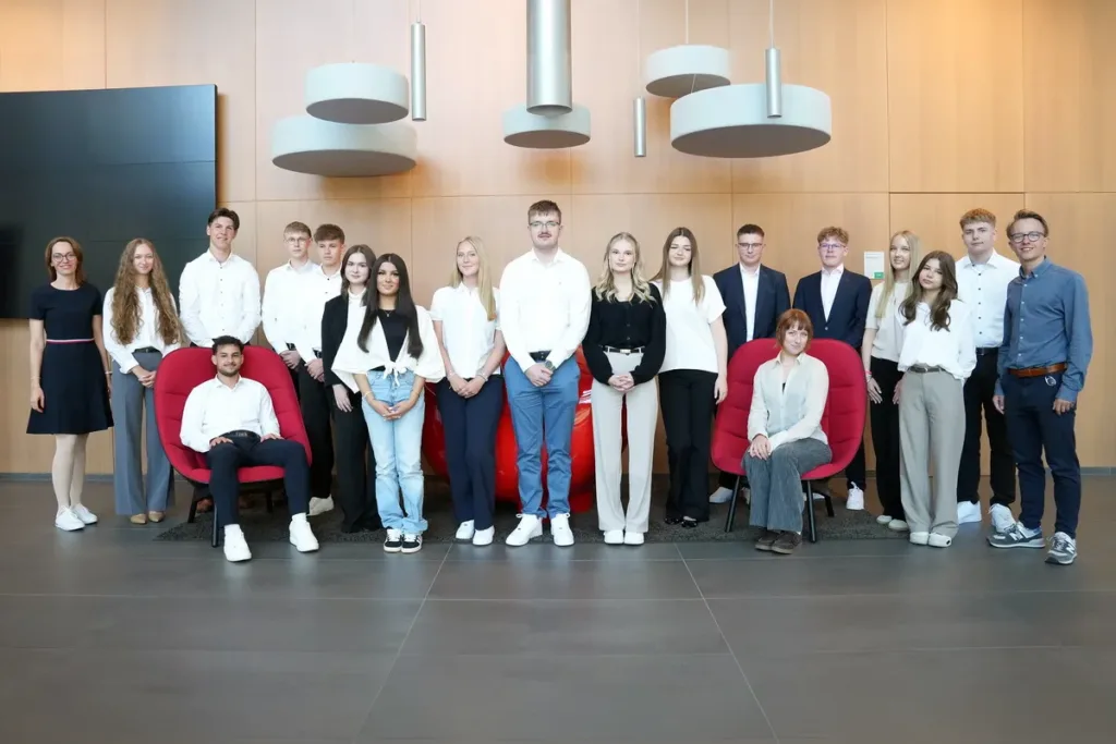 Group of students and two teachers posing in a modern lobby with red chairs and wooden panel walls.