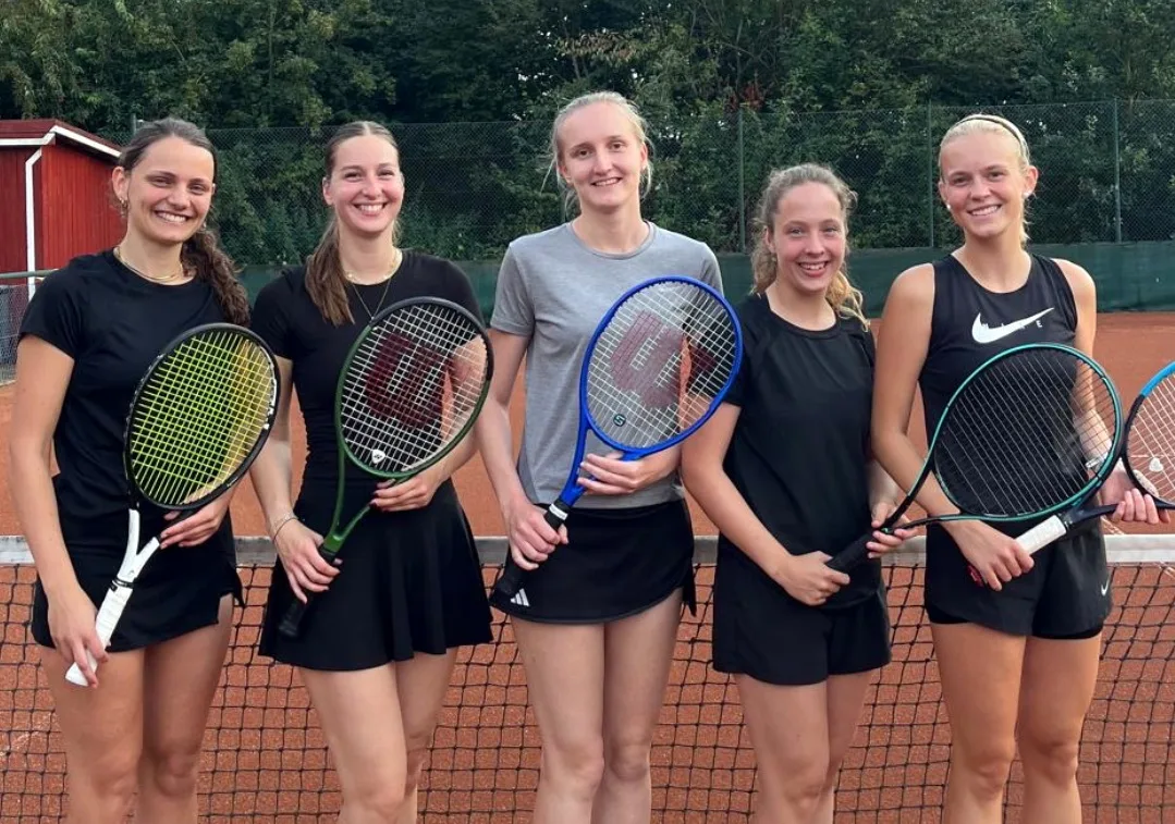 Five young women stand side by side on a clay tennis court, each holding a racket and smiling at the camera.