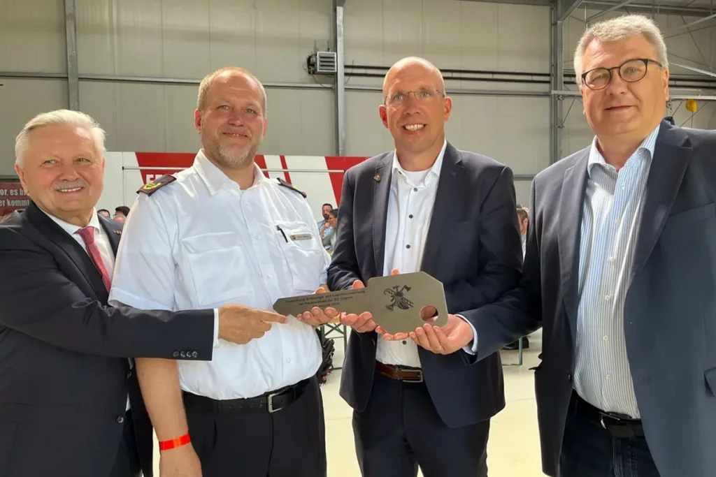 Four men in business and uniform attire pose in an industrial setting, presenting a large ceremonial metal key to mark a milestone.