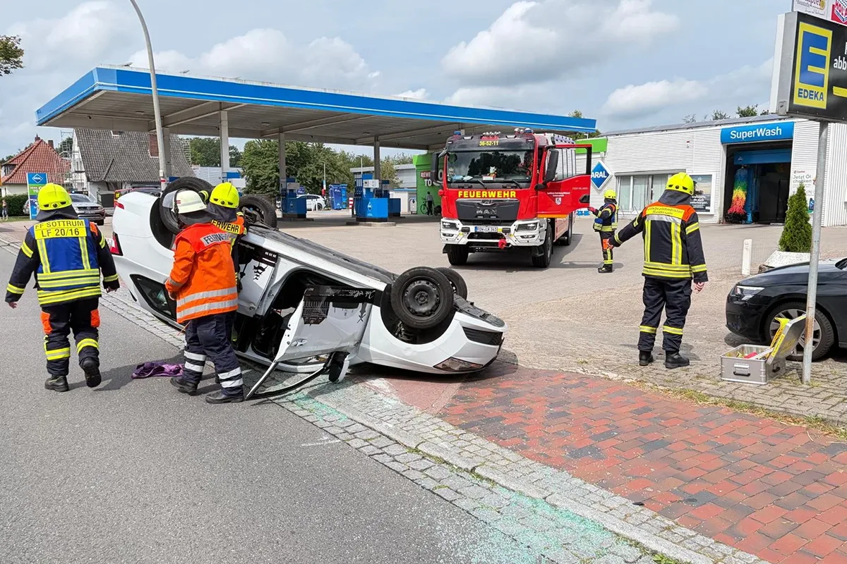 White car is overturned on its roof at a gas station, firefighters and a red fire engine in the background aid at the scene.