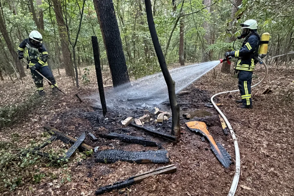 Firefighters in protective gear spray water on charred debris in a wooded area to extinguish a small brush fire.