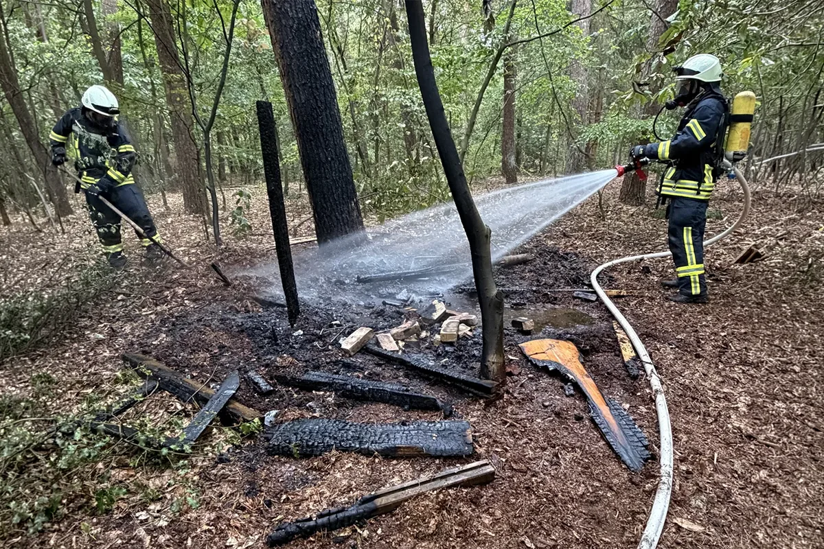 Firefighters in protective gear spray water on charred debris in a wooded area to extinguish a small brush fire.