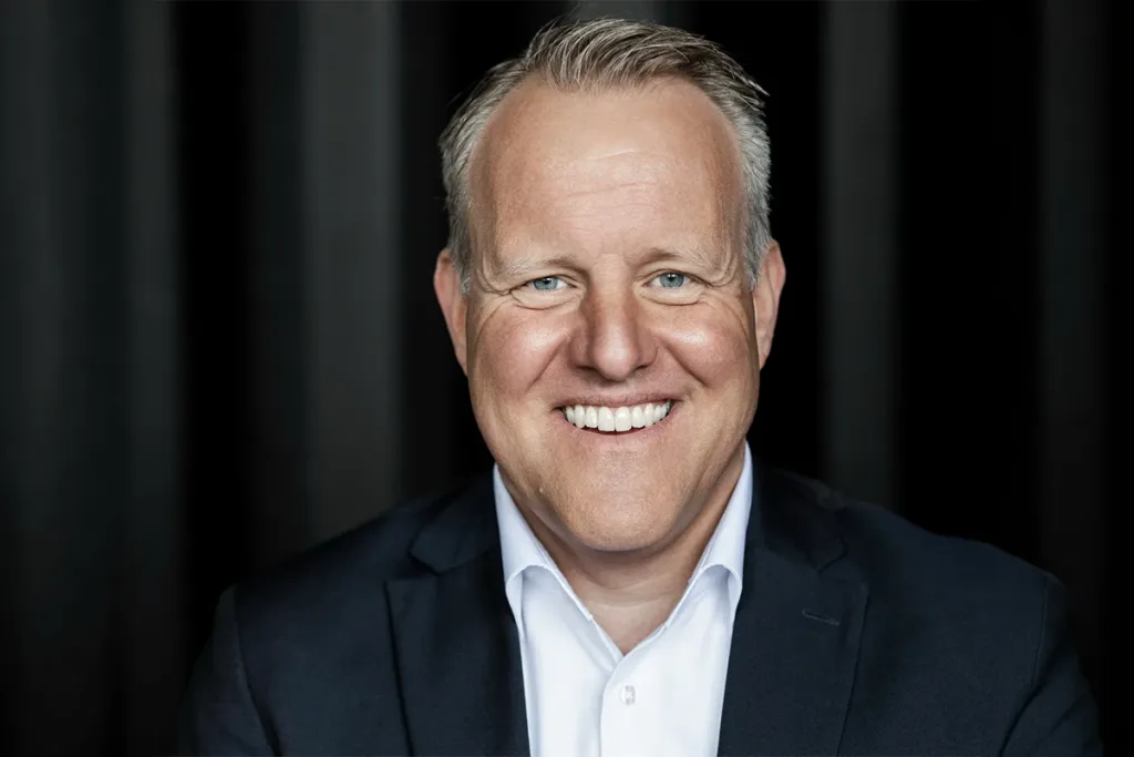 Smiling man in a dark blazer and white shirt against a black backdrop, looking at the camera.