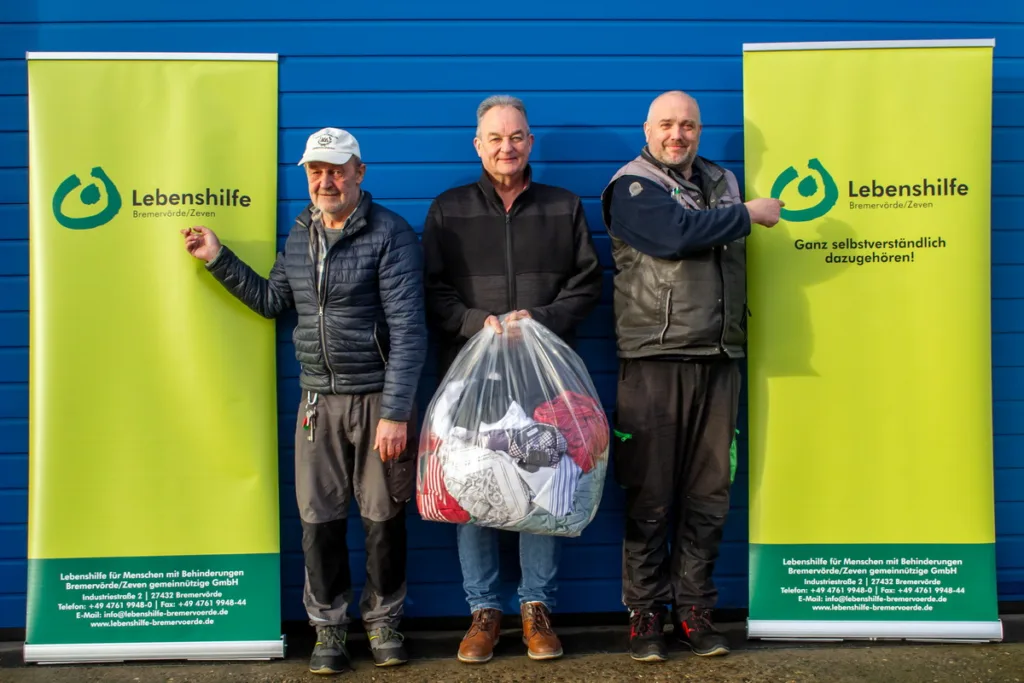Three men stand in front of a blue wall between Lebenshilfe banners; the center man holds a large bag of donated clothes.