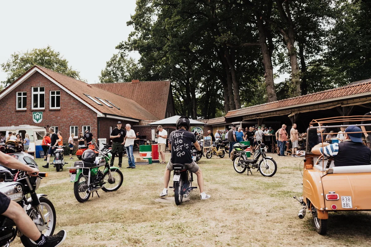 Outdoor motorcycle gathering on a grassy field with vintage bikes and a brick building in the background.