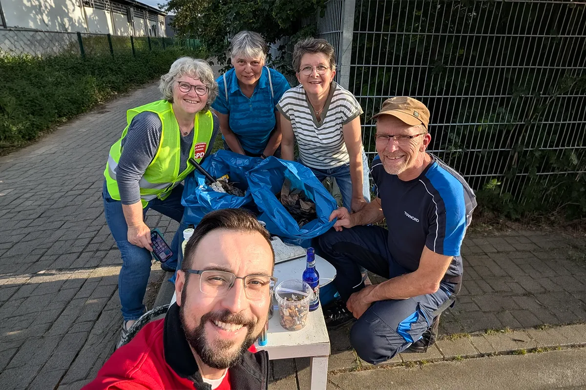 Five volunteers on a sidewalk pose for a selfie beside a full blue trash bag, with bottles and a small table nearby after a community cleanup.