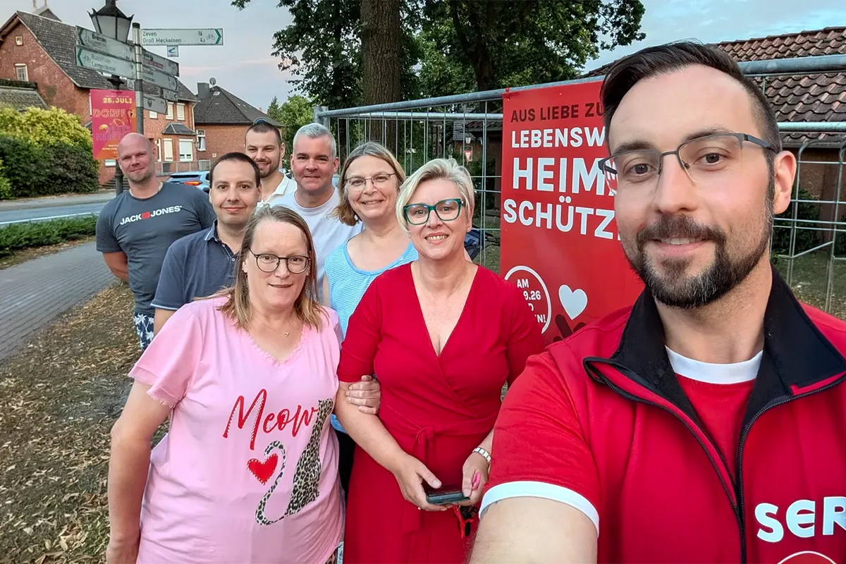 Eight adults pose for a group selfie outdoors beside a metal fence and a red banner, smiling at the camera.