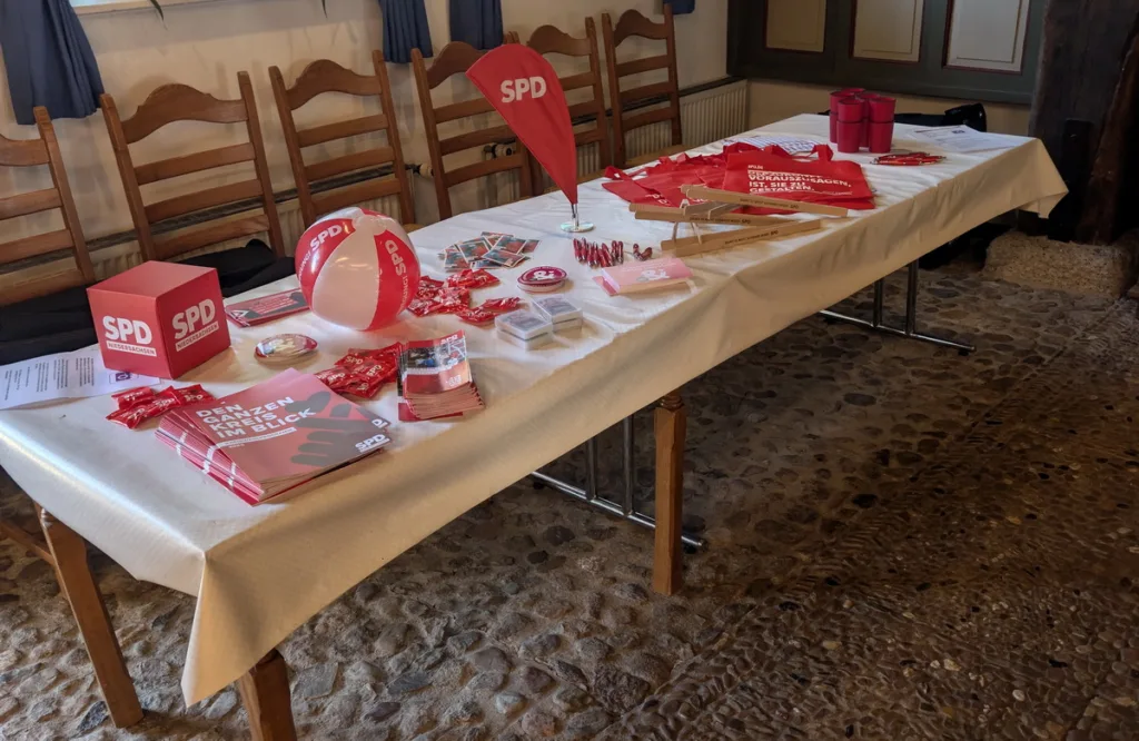 Table covered with a white cloth showcasing SPD campaign materials: red brochures, a red-and-white SPD balloon, stickers, and a pink SPD cube.