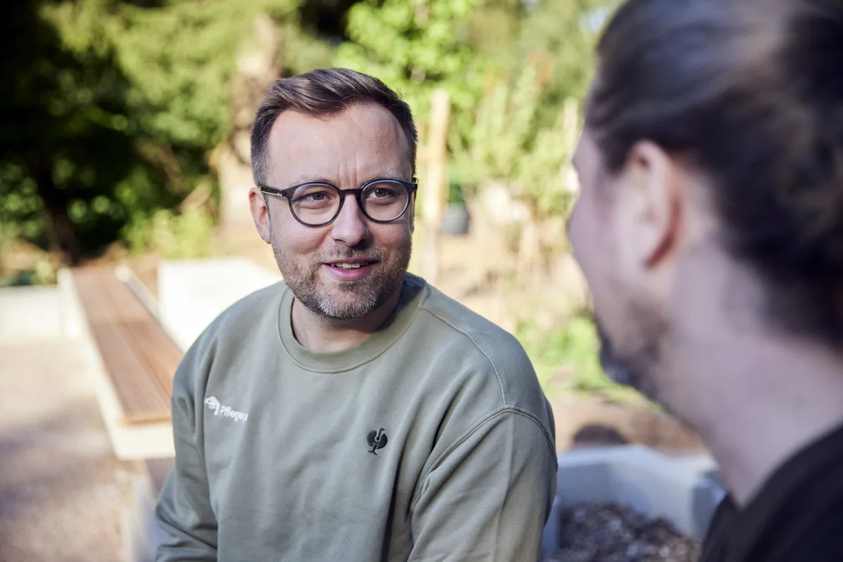 Man with glasses and stubble wearing an olive green sweatshirt talks with another person outdoors in a sunny garden.