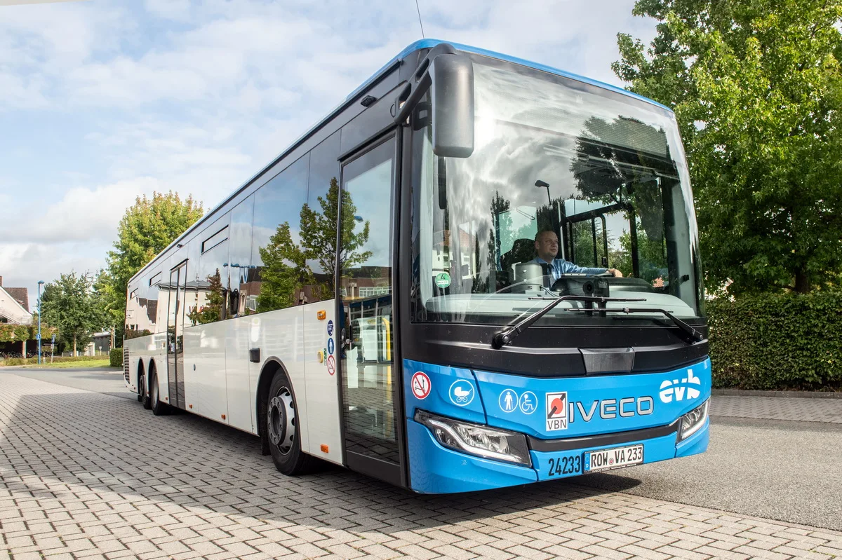 Blue Iveco city bus with driver visible in the cab, parked on a cobblestone street with trees in the background.