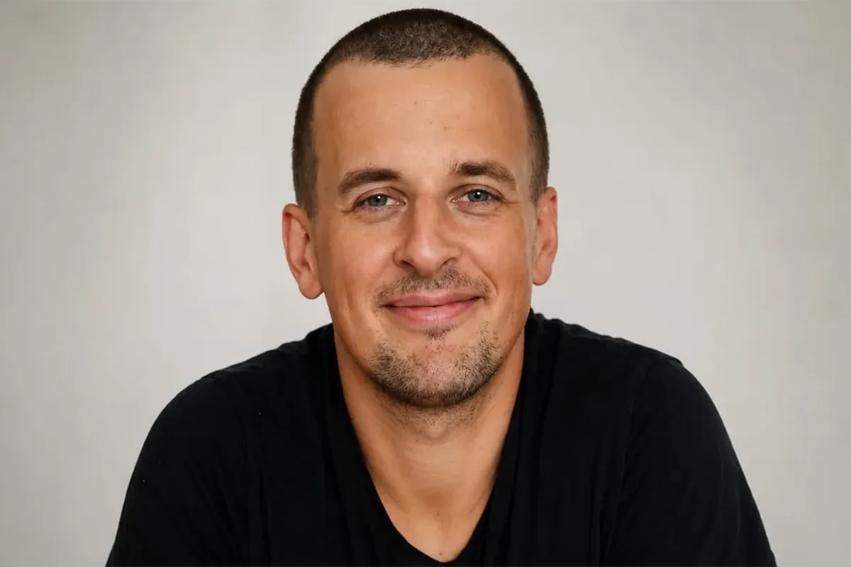 Smiling man with short hair and light stubble, wearing a black shirt, against a neutral gray background (head-and-shoulders portrait).