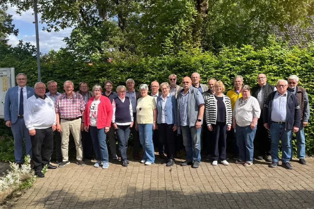 Group of about two dozen adults standing in a line outdoors, posing for a photo in front of a hedge and trees on a sunny day, casually dressed.