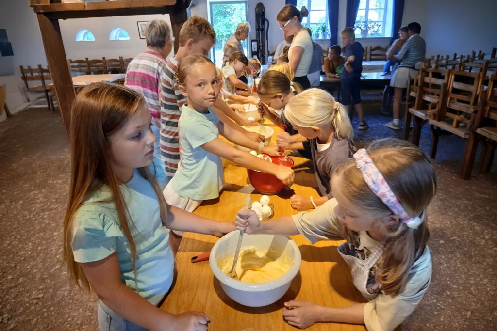 Children stand along a long wooden table, stirring batter in bowls as part of a group cooking activity in a classroom/kitchen setting.