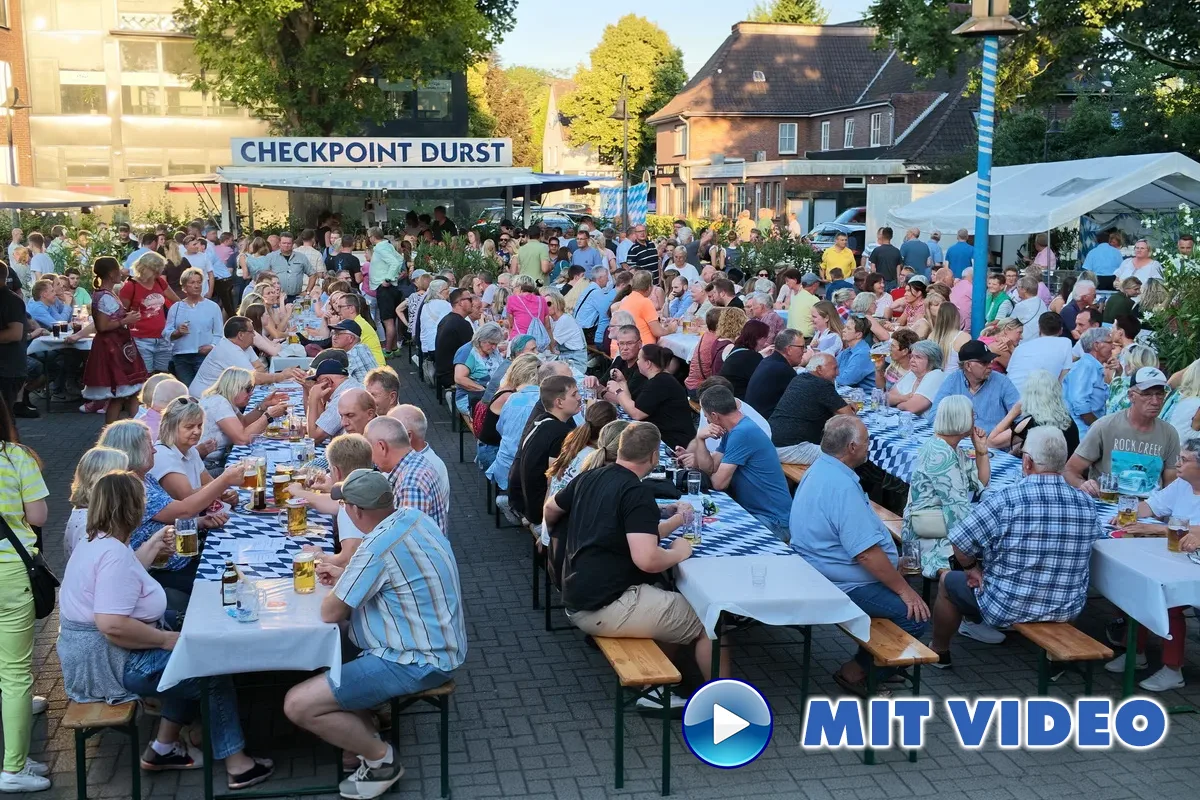 Crowd of people at outdoor beer festival, seated at long communal tables under a 'CHECKPOINT DURST' sign, enjoying food and drinks on a sunny day.