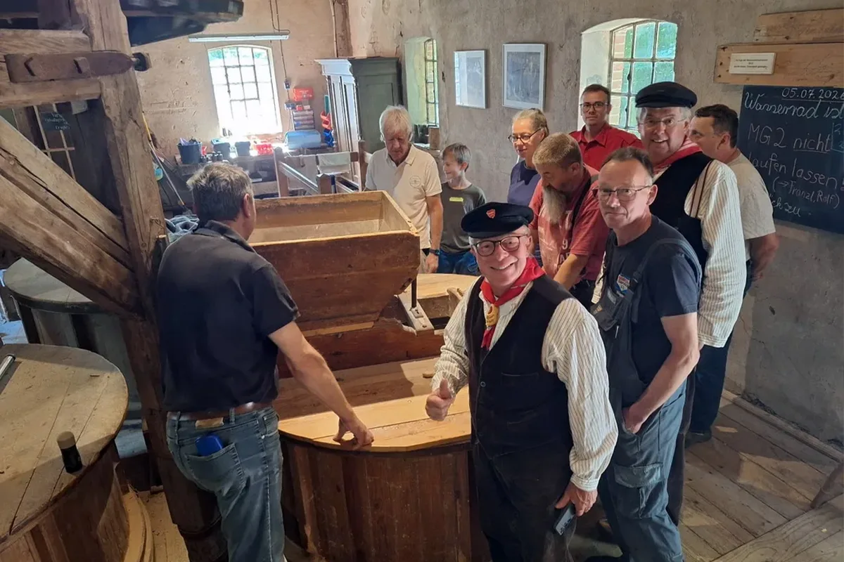 Group of people in a rustic workshop watching a large wooden milling device being operated by a man in a dark shirt at the front.
