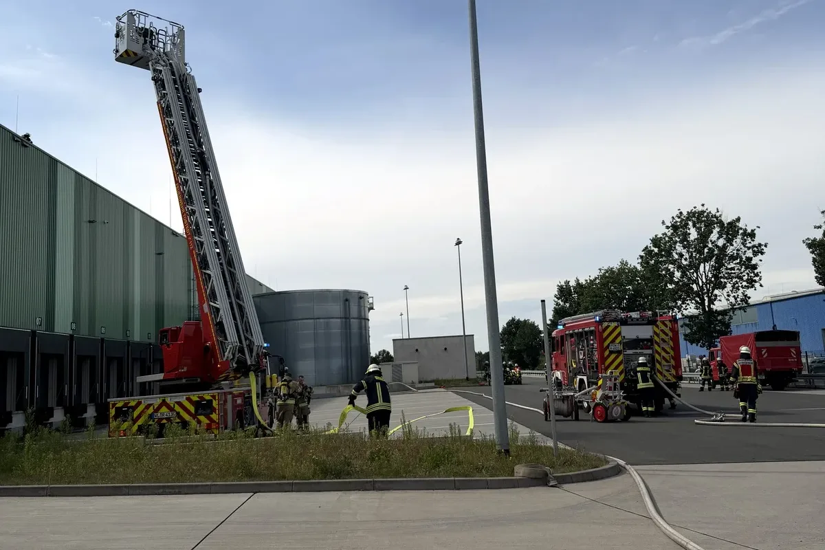 Firefighters deploy a large aerial ladder near a green industrial building, with hoses laid out and multiple emergency vehicles on scene.