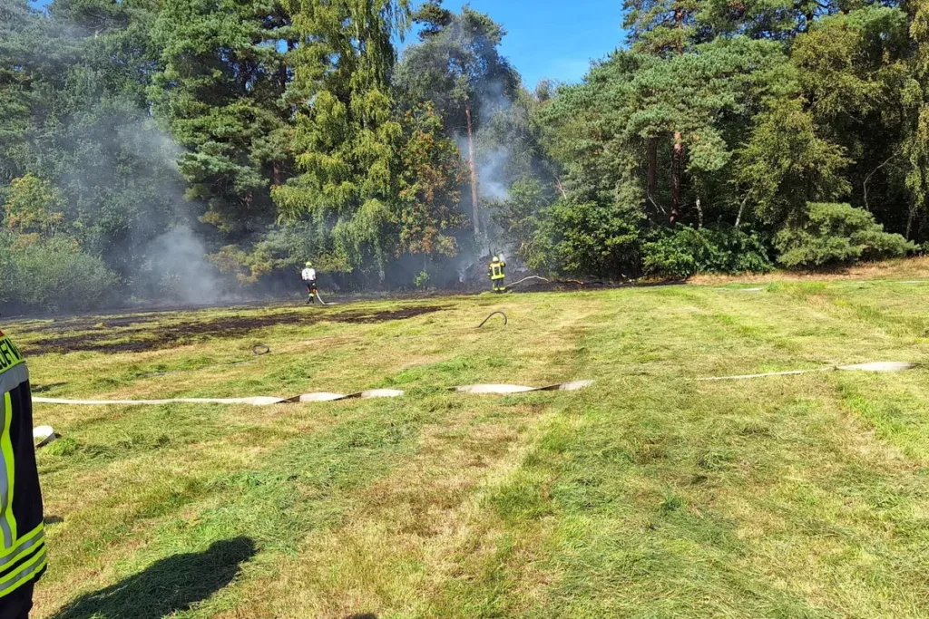 Firefighters in yellow gear extinguish a smoldering grass fire in a field with smoke and trees in the background.