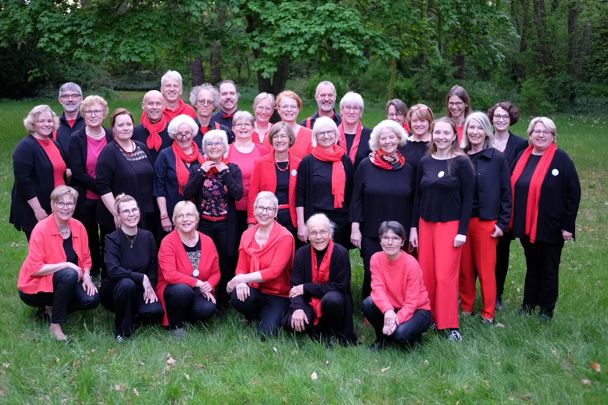 Group of about 30 adults posing outdoors in a park, many wearing red and black outfits and scarves, smiling at the camera.