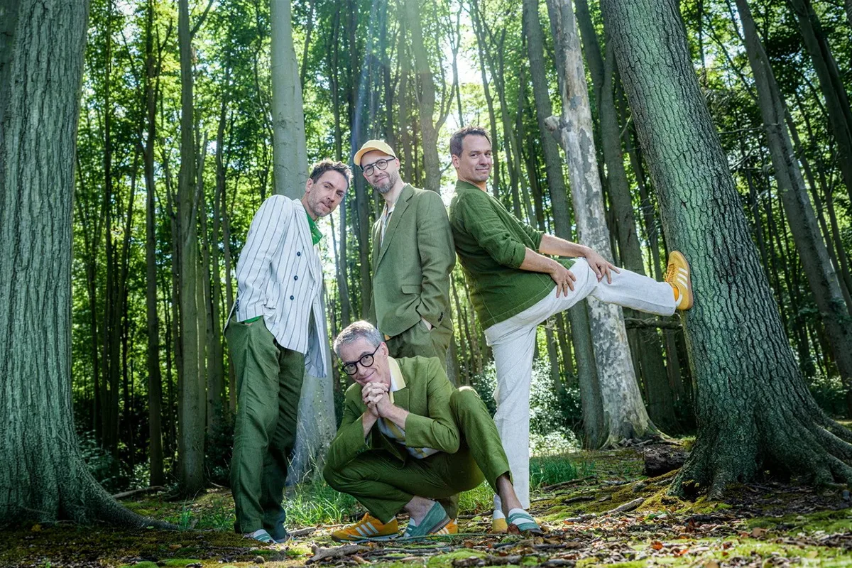 Four men in green outfits posing playfully in a sunlit forest among tall trees, one with a leg kicked against a tree trunk.