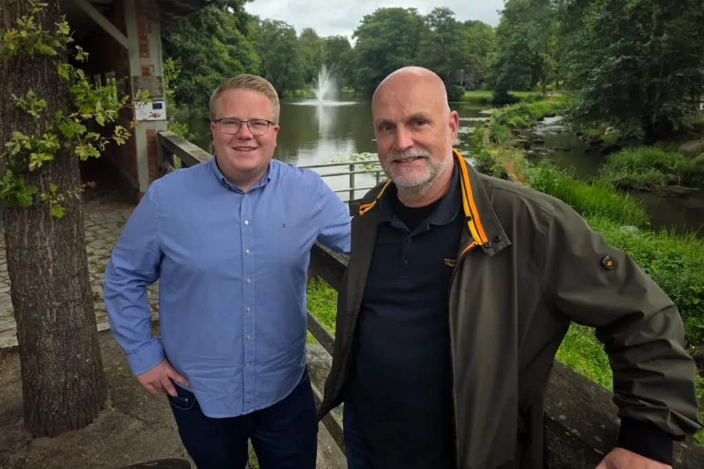 Two men posing outdoors by a pond with a fountain, smiling at the camera.