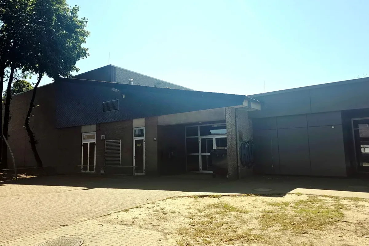 Modern brick building with a covered entrance and a parked car, trees on the left, under a bright blue sky.
